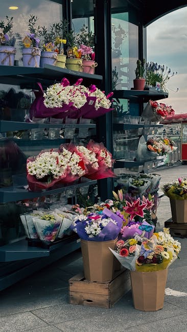 An outdoor floral display at a flower shop features multiple bouquets arranged on metal shelving and in brown paper bags on the ground. The bouquets include vibrant pink, white, purple, and yellow flo